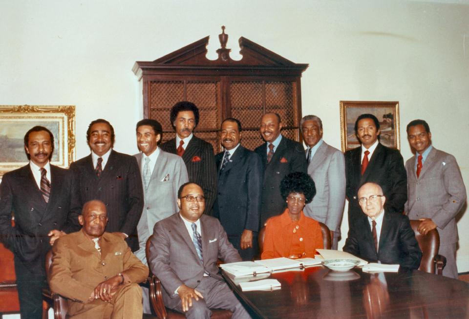Photo of 9 Black men standing, wearing suits, 3 Black men and one Black woman seated, dressed professionally.